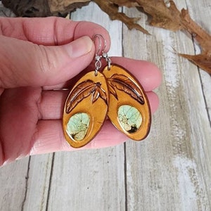May include: A pair of brown leather earrings with a green and white stone inlay and a carved feather design.