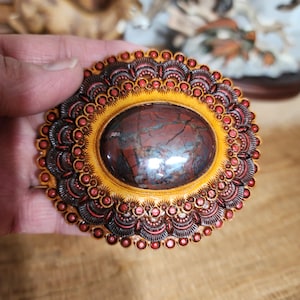 May include: Oval-shaped brooch with a polished, reddish-brown stone center. The stone is set in a yellow frame, surrounded by a detailed, ornate border of red and dark brown accents. The brooch is held in a hand, showcasing its intricate design.