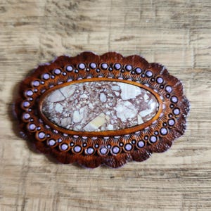 May include: Oval-shaped, handcrafted leather belt buckle with a central stone inlay. The buckle features a brown leather border with a scalloped edge and small purple accents. The stone has a mottled pattern of white, brown, and beige.