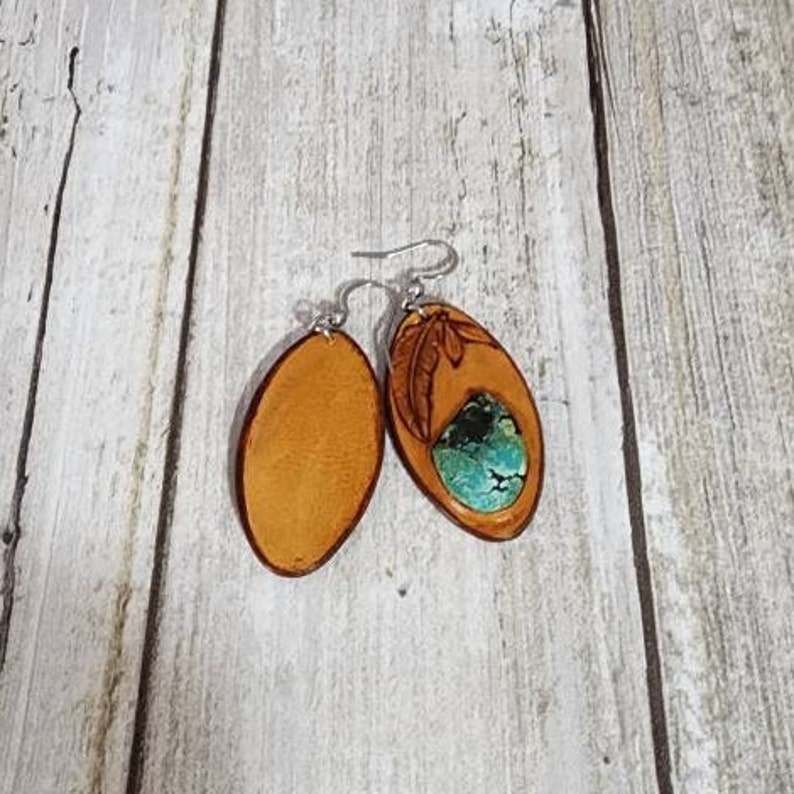 May include: A pair of oval-shaped leather earrings with a turquoise stone inlay. One earring has a carved feather design, while the other is plain.