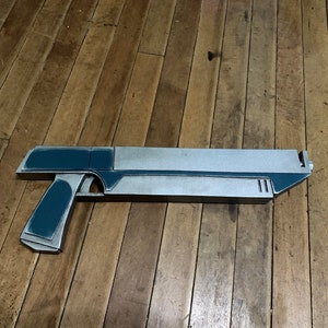 Puede incluir: Una pistola de juguete futurista de color plateado y verde azulado. La pistola tiene un cañón largo y una empuñadura de pistola.