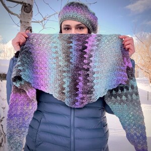 May include: A handmade crochet shawl and matching beret in shades of purple, blue, and gray. The shawl has a scalloped edge and a chevron pattern. The beret is a solid color with a subtle gradient. The items are displayed outdoors.