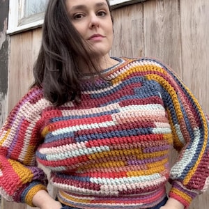 May include: A colorful, handmade crochet sweater with a round neckline. The sweater features a geometric pattern with stripes of red, blue, yellow, and pink. The sleeves are long and the fit is relaxed. The sweater is a cozy and stylish garment.