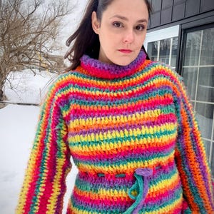 May include: A handmade, rainbow-striped crochet sweater with a high neck and long sleeves. The sweater features a cinched waist with a drawstring. The color palette includes vibrant hues of pink, yellow, orange, green, and purple, creating a bold and colorful design.