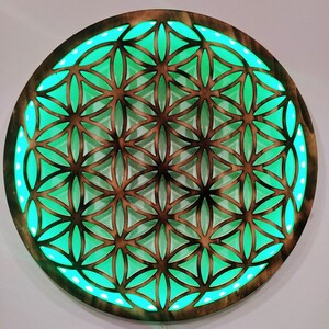 May include: A round wooden wall decor with a geometric pattern of a flower of life design. The pattern is cut out of the wood and illuminated with green LED lights.