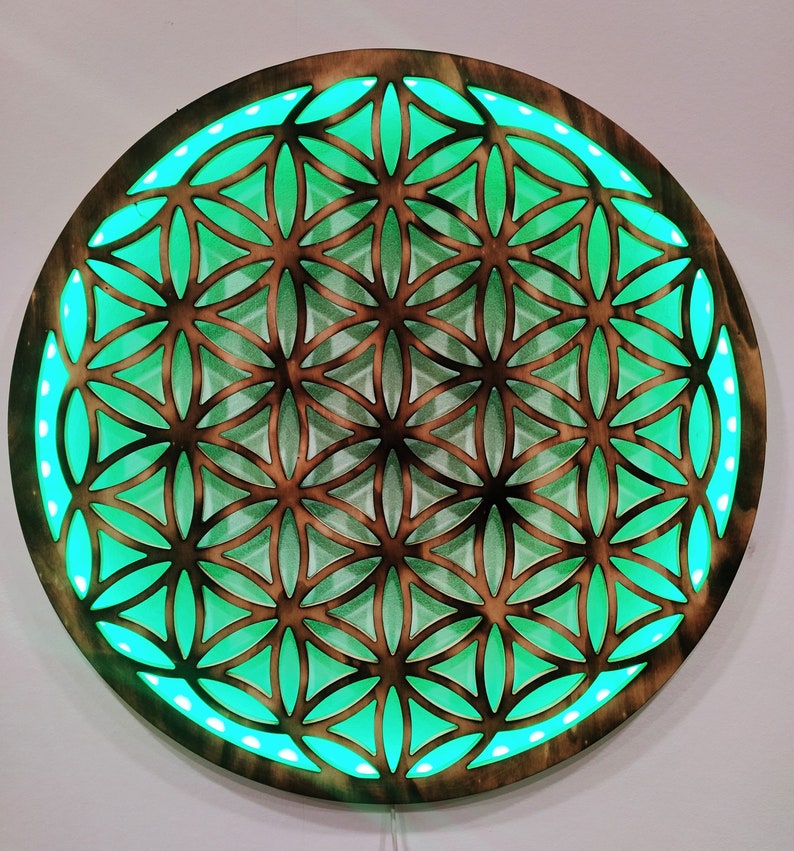 May include: A round wooden wall decor with a geometric pattern of a flower of life design. The pattern is cut out of the wood and illuminated with green LED lights.