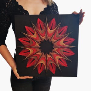 Könnte beinhalten: Ein quadratisches schwarzes Brett mit einem geometrischen Fadenkunst-Design. Das Muster besteht aus leuchtend roten und goldbeigen Fäden, die um kleine Nägel gewickelt sind und eine symmetrische, sternförmige Mandala-Form mit leerer Mitte bilden.