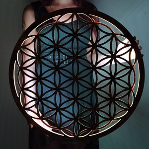 May include: A wooden wall hanging in the shape of a flower of life. The intricate design is cut out of the wood, creating a geometric pattern. The wood is a dark brown color and the design is illuminated from behind, creating a warm glow.