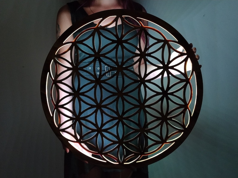 May include: A wooden wall hanging in the shape of a flower of life. The intricate design is cut out of the wood, creating a geometric pattern. The wood is a dark brown color and the design is illuminated from behind, creating a warm glow.