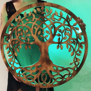May include: A wooden tree of life wall decor with a circular shape and intricate carvings. The wood is a dark brown color and the background is a bright teal green.