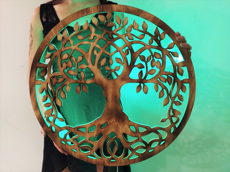 May include: A wooden tree of life wall decor with a circular shape and intricate carvings. The wood is a dark brown color and the background is a bright teal green.