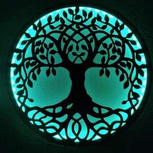 May include: A circular wooden wall decor with a cutout design of a tree of life. The tree is black and the background is a teal blue color.