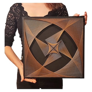 Op de afbeelding: Een vierkant draadkunstwerk met een zwarte achtergrond en complexe geometrische patronen, gevormd door dunne beige, bruine en gouden draden gespannen tussen spijkers langs de randen, wat een centrale vierpuntige ster vormt.