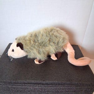 May include: A plush opossum toy with a light brown and beige color scheme. The toy has a long, pink tail and a furry body. The opossum is lying on a dark gray surface.