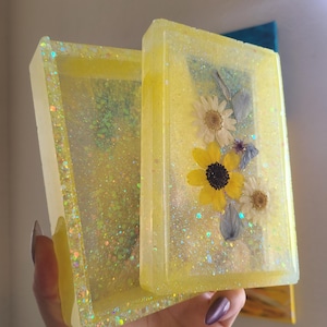 May include: Two rectangular, yellow resin trays with glitter and dried flowers. One tray is open, revealing the interior. The flowers include yellow sunflowers and white daisies. The trays are handmade and decorative.