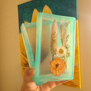 May include: A rectangular, clear resin tray with a light teal border, holding dried flowers. The flowers include a peach rose, white daisy, and tall, dried grasses. The tray is held in front of a painting of a sunflower.