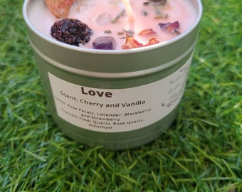 May include: A green candle jar with a pink wax top and crystals embedded in the wax. The jar has a label that reads "Love" and "Scent: Cherry and Vanilla". The label also lists the ingredients: Rose Petals, Lavender, Blackberry, and Strawberry. The crystals are Clear Quartz, Rose Quartz, and Amethyst.