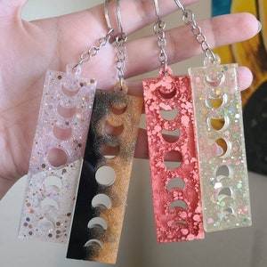 May include: Four keychains with different colored resin and glitter. Each keychain features a silver chain and a rectangular charm with a crescent moon design.