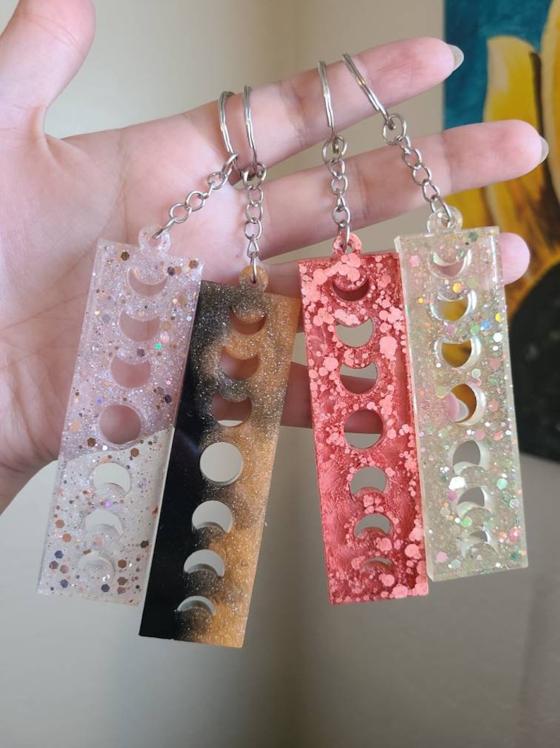 May include: Four keychains with different colored resin and glitter. Each keychain features a silver chain and a rectangular charm with a crescent moon design.
