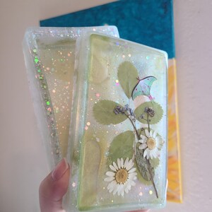 May include: Two rectangular resin coasters, one open and one closed, with dried flowers, leaves, and a moth design. The coasters are filled with glitter and have a light green hue. The background features a blue and yellow abstract painting.