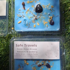 K&ouml;nnte beinhalten: Ein blaues Wachs-Melt mit dem Text "Safe Travels" und dem Duft "Ocean Breeze, Awapuhi, & Seaberry". Kr&auml;uter: Ingwer, Nelke und Thymian. Kristalle: Schwarzer Onyx, klarer Quarz und Granat. Das Wachs-Melt befindet sich in einem durchsichtigen Plastikbeh&auml;lter.