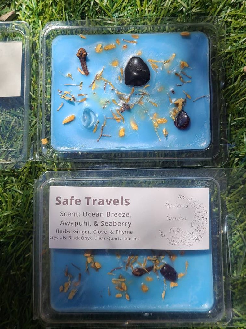 K&ouml;nnte beinhalten: Ein blaues Wachs-Melt mit dem Text "Safe Travels" und dem Duft "Ocean Breeze, Awapuhi, & Seaberry". Kr&auml;uter: Ingwer, Nelke und Thymian. Kristalle: Schwarzer Onyx, klarer Quarz und Granat. Das Wachs-Melt befindet sich in einem durchsichtigen Plastikbeh&auml;lter.