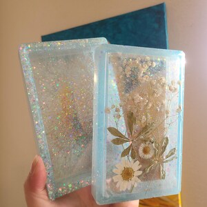 May include: Two rectangular resin art pieces. One contains glitter, the other features dried white flowers and leaves. Both are encased in light blue resin with glitter accents. The pieces are held in a hand.