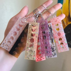 May include: A set of six colorful moon phase keychains. Each keychain is made of clear resin with glitter and features a different color and design. The keychains are attached to a silver key ring.
