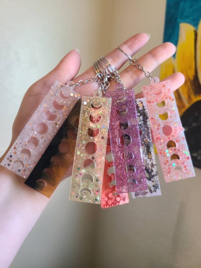 May include: A set of six colorful moon phase keychains. Each keychain is made of clear resin with glitter and features a different color and design. The keychains are attached to a silver key ring.