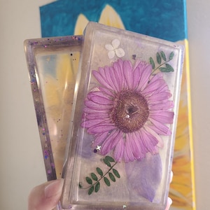 May include: Rectangular clear resin case with a pressed purple flower, leaves, and glitter. The case is open, revealing a second case. The background includes a painting of a sunflower. The case is approximately 10 cm long.