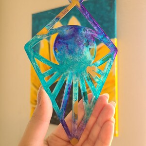 May include: A decorative, abstract resin ornament in shades of teal, purple, and gold. The ornament has a diamond shape with a circular center and radiating lines. The background features a painting with yellow and teal colors.