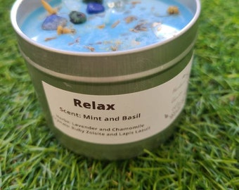 Pode incluir: Um frasco de vela de metal verde com uma vela de cera azul dentro. A vela tem pequenos cristais e flores secas embutidos na cera. O rótulo no frasco diz "Relax" e "Scent: Mint and Basil". Ervas: Lavanda e camomila. Cristais: Zoisita rubi e lapis-lazúli.