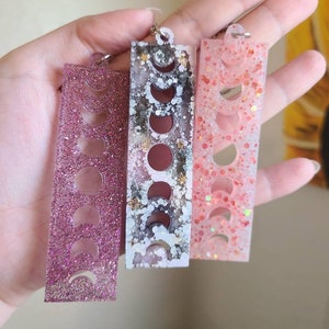 May include: Three rectangular resin keychains with a moon phase design. The keychains are pink, white with black and gold accents, and peach with iridescent glitter.