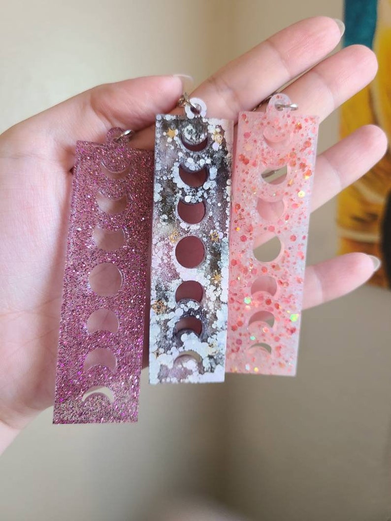 May include: Three rectangular resin keychains with a moon phase design. The keychains are pink, white with black and gold accents, and peach with iridescent glitter.