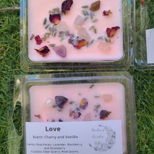 K&ouml;nnte beinhalten: Eine rosa Sojawachskerze mit einem floralen und kristallinen Design. Die Kerze ist mit "Love" beschriftet und hat einen Duft nach Kirsche und Vanille. Die verwendeten Kr&auml;uter sind Rosenbl&auml;tter, Lavendel, Brombeere und Erdbeere. Die verwendeten Kristalle sind klarer Quarz, Rosenquarz und Amethyst.