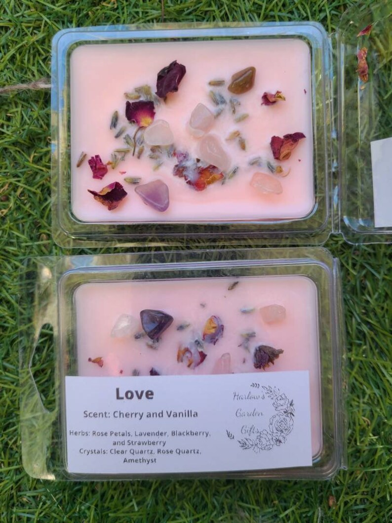 K&ouml;nnte beinhalten: Eine rosa Sojawachskerze mit einem floralen und kristallinen Design. Die Kerze ist mit "Love" beschriftet und hat einen Duft nach Kirsche und Vanille. Die verwendeten Kr&auml;uter sind Rosenbl&auml;tter, Lavendel, Brombeere und Erdbeere. Die verwendeten Kristalle sind klarer Quarz, Rosenquarz und Amethyst.