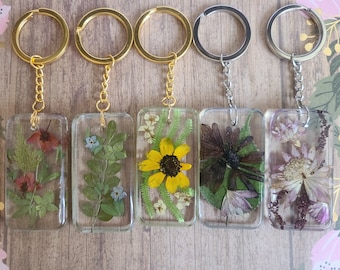 May include: Five rectangular keychains with gold or silver-colored hardware. Each keychain features dried flowers encased in clear resin. The flowers include red, green, yellow, and purple hues, offering a natural, botanical aesthetic.