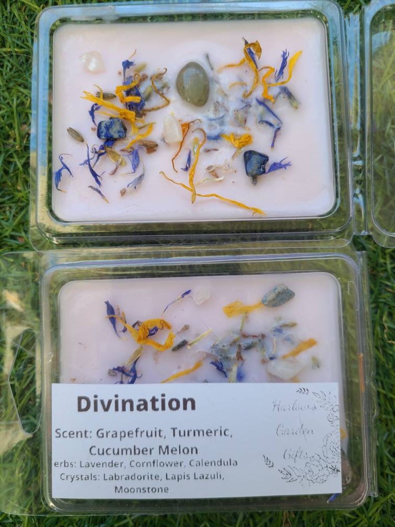 K&ouml;nnte beinhalten: Eine rosa Sojawachskerze mit getrockneten Blumen und Kristallen. Die Kerze ist mit "Divination" beschriftet und listet den Duft als "Grapefruit, Kurkuma, Gurkenmelone" auf. Das Etikett listet auch die verwendeten Kr&auml;uter auf: Lavendel, Kornblume, Ringelblume. Die verwendeten Kristalle sind Labradorit, Lapislazuli und Mondstein.
