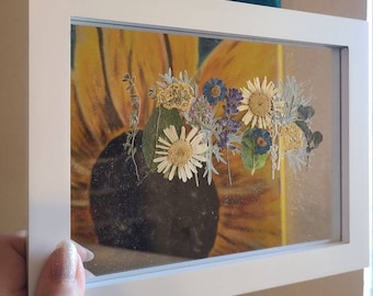 Könnte beinhalten: Ein weißer Rahmen mit einer Glasfront, in dem ein gepresstes Blumenarrangement enthalten ist. Die Blumen sind eine Mischung aus Weiß, Gelb und Lila. Der Hintergrund ist ein Gemälde einer Sonnenblume.