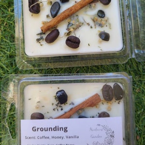 K&ouml;nnte beinhalten: Eine wei&szlig;e Kerze mit einem erdigen Duft, die Kaffeebohnen, Zimtstangen und schwarze Onyx-Kristalle enth&auml;lt. Das Etikett lautet "Grounding" und listet den Duft als "Kaffee, Honig, Vanille" und die Kr&auml;uter als "Brennnessel, Salbei, Kaffeebohnen, Zimt" auf. Das Etikett gibt auch "Kristalle: Schwarzer Onyx" und "Harlours Garden Gifts" an.