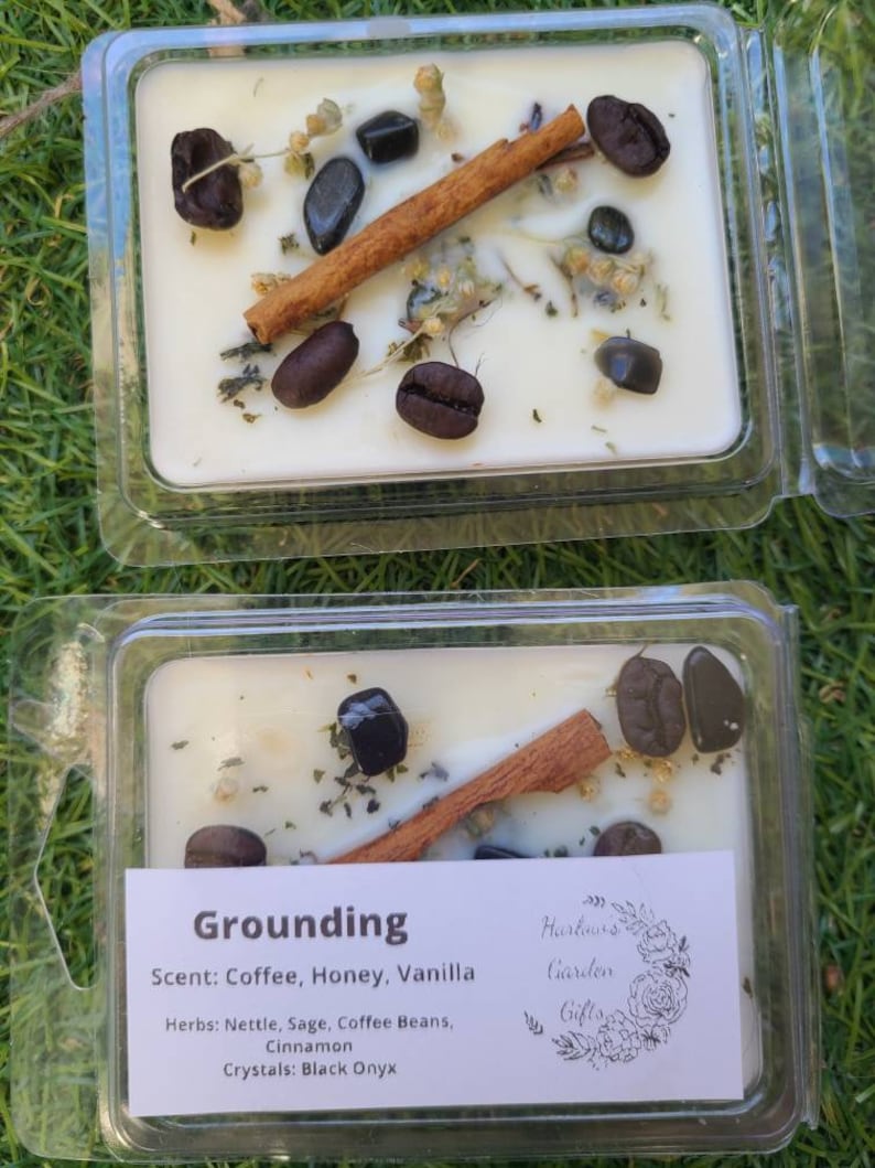 K&ouml;nnte beinhalten: Eine wei&szlig;e Kerze mit einem erdigen Duft, die Kaffeebohnen, Zimtstangen und schwarze Onyx-Kristalle enth&auml;lt. Das Etikett lautet "Grounding" und listet den Duft als "Kaffee, Honig, Vanille" und die Kr&auml;uter als "Brennnessel, Salbei, Kaffeebohnen, Zimt" auf. Das Etikett gibt auch "Kristalle: Schwarzer Onyx" und "Harlours Garden Gifts" an.