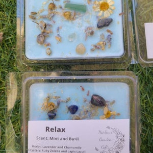 K&ouml;nnte beinhalten: Ein blaues Wachs-Melt mit getrockneten Blumen und Kristallen. Das Etikett lautet "Relax" mit dem Duft "Minze und Basilikum", Kr&auml;utern "Lavendel und Kamille" und Kristallen "Rubinzoisit und Lapislazuli". Das Etikett enth&auml;lt auch den Text "Harlows Garden Gifts".