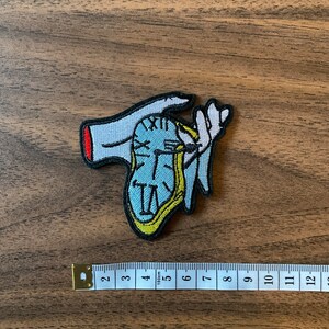 May include: An embroidered patch featuring two hands holding a clock face. The clock face is light blue with black Roman numerals and black hands, and is outlined in yellow.