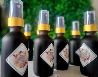 May include: A row of six black glass spray bottles with gold lids and white labels. The labels have a diamond shape and feature the text "White Sage Lavender Rose 1 oz".