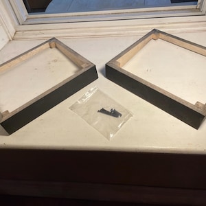 Peut inclure: Deux cadres rectangulaires en bois avec une finition extérieure teintée foncée et des bords intérieurs en bois naturel. Un petit sachet en plastique transparent contenant plusieurs vis métalliques est placé entre les deux cadres pour l'assemblage.