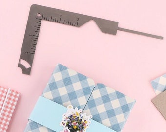 Puede incluir: Una escuadra de acero inoxidable con medidas en centímetros se coloca sobre un regalo envuelto en papel a cuadros azul y blanco. Una cinta azul claro con una pegatina floral envuelve el regalo. Se ve un cúter.