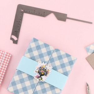May include: A stainless steel angle ruler with inch measurements is positioned above a gift wrapped in blue and white checkered paper. A light blue band with a floral sticker wraps the gift. A craft knife is visible.