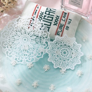 May include: Two white, intricately cut paper doilies with a floral design are displayed on a light blue surface. A small, rectangular fabric item with the words "SUGAR PURE CANE" is in the background. The doilies are delicate and detailed.