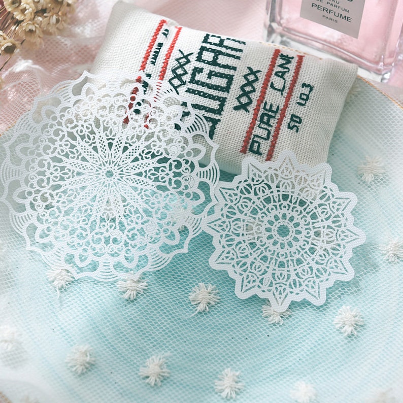 May include: Two white, intricately cut paper doilies with a floral design are displayed on a light blue surface. A small, rectangular fabric item with the words "SUGAR PURE CANE" is in the background. The doilies are delicate and detailed.