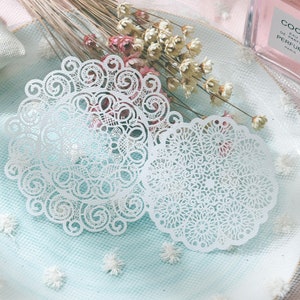 May include: Two white, intricately designed paper doilies with a delicate, lace-like pattern. The doilies are circular with scalloped edges. Dried flowers and a perfume bottle are in the background, suggesting a vintage or craft theme.